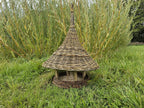 Handwoven willow bird feeder with a conical roof standing on the lawn in front of tall ornamental grasses in late summer