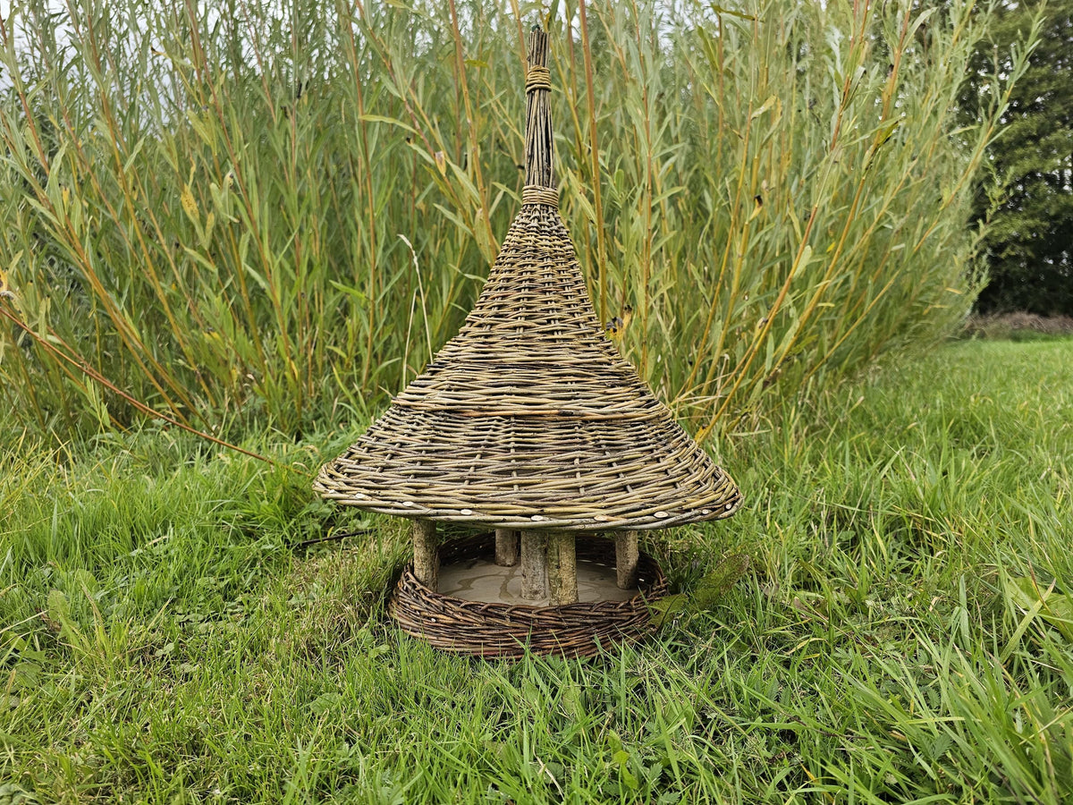 Handwoven willow bird feeder with a conical roof standing on the lawn in front of tall ornamental grasses in late summer