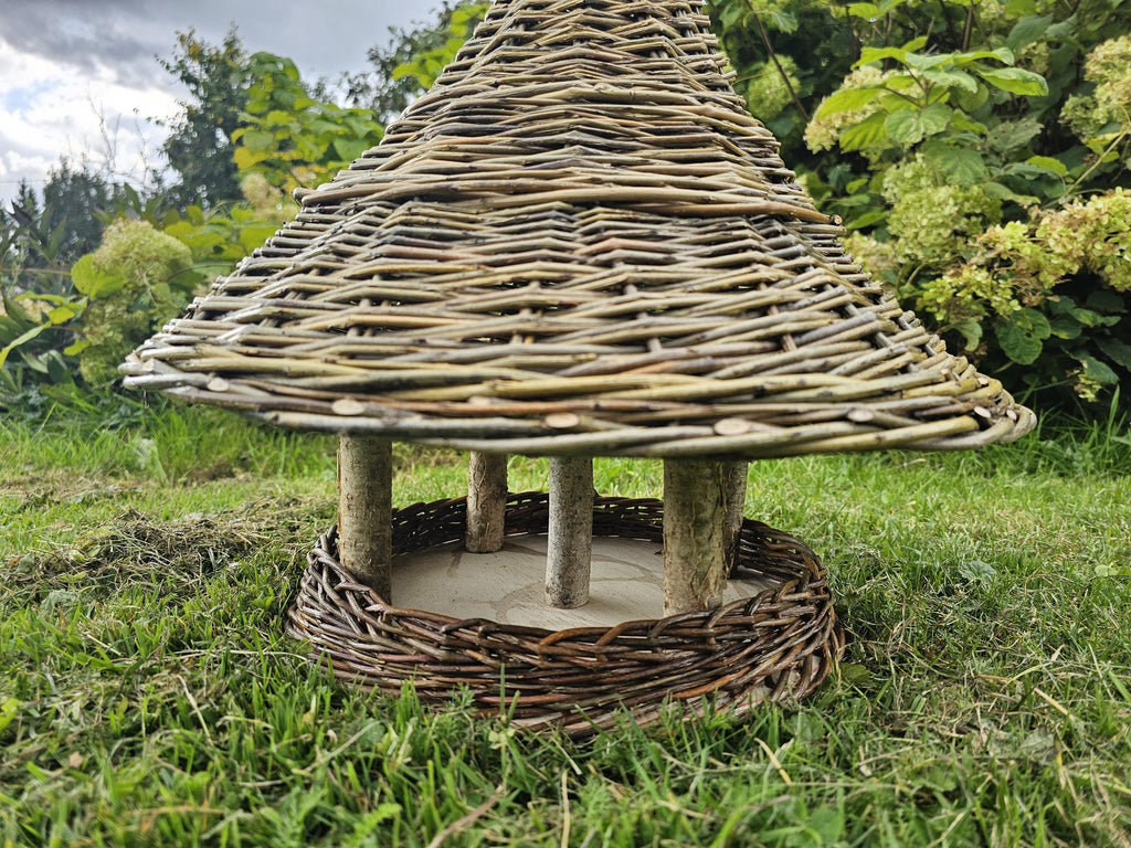 Close view of the willow bird table showing the wide woven roof resting on branch posts above the wooden seed platform with its woven rim