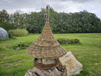 Willow garden bird feeder with a tall pointed roof set on a log in an open meadow with trees and a greenhouse in the background