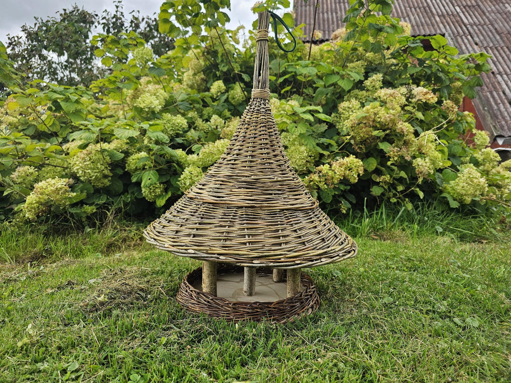 Large handwoven bird table with a wide conical willow roof on branch posts above the seed tray, on the grass by a flowering shrub