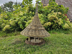 Large handwoven bird table with a wide conical willow roof on branch posts above the seed tray, on the grass by a flowering shrub