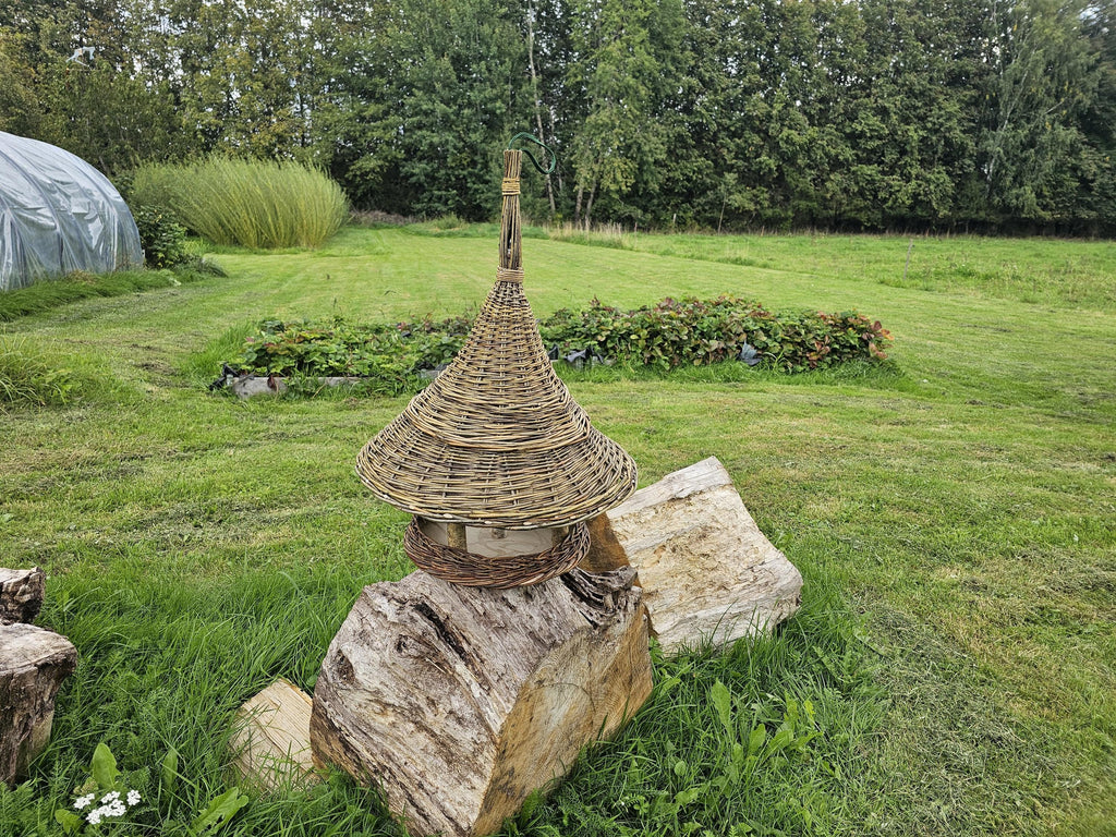 Hanging willow bird table with a conical roof placed on split logs in a mown meadow, showing its full height and woven tray rim