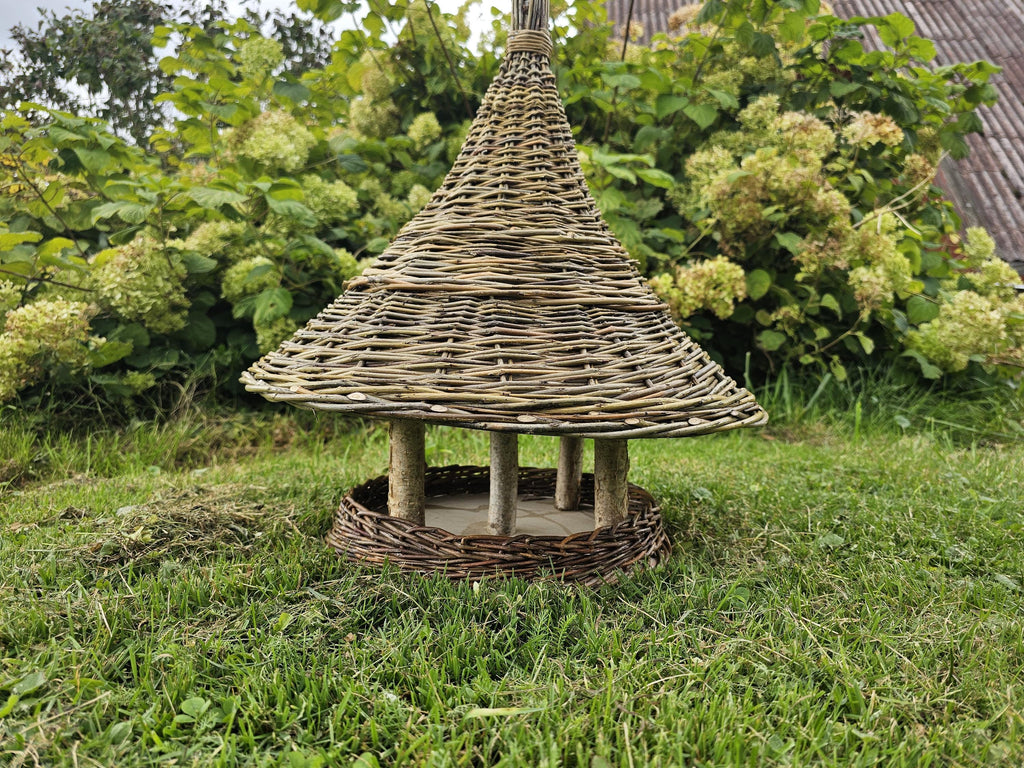 Side view of the willow bird table with the wide woven roof raised on wooden branch posts above the round seed platform, on the lawn