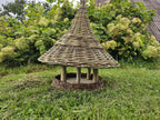 Side view of the willow bird table with the wide woven roof raised on wooden branch posts above the round seed platform, on the lawn