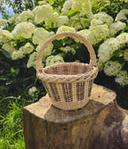 Mini round wicker basket with a single arched handle on a tree stump before white hydrangeas, its tapered body worked in a tight uniform weave beneath a braided rim.