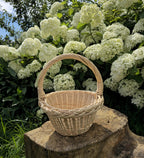 Round handwoven willow basket with a tapered body, circular base and braided rim on a tree stump by hydrangeas, woven from thin flexible rods with an arched handle.