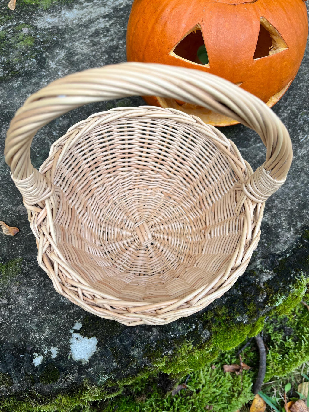 Top view of the round willow basket showing the deep open interior and spiral-woven base, placed in front of a carved pumpkin for a seasonal display.