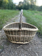 Front view of the willow market basket from the nesting set on a gravel path, the woven band around the lower body and the handle collar visible.