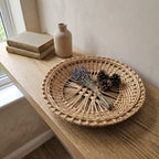 Openwork wicker pastry display tray with a lattice centre and plaited rim on a window ledge, holding a bundle of dried lavender and pine cones.