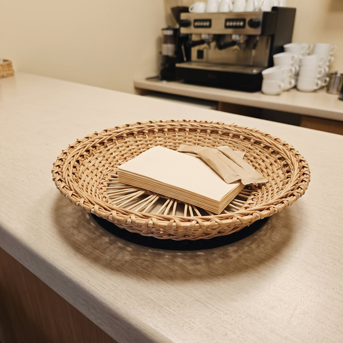 Lace-weave willow tray holding paper napkins on a café counter next to a coffee machine and stacked white cups.
