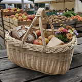 Großer ovaler Marktkorb mit zwei Henkeln, darin Sauerteigbrot, Äpfel und ein kleiner Blumenstrauß, an einem Stand auf dem Bauernmarkt