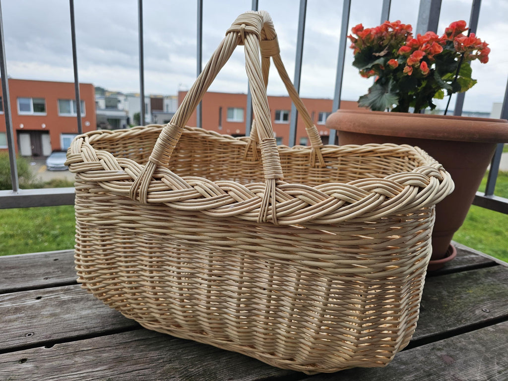 Deep oval wicker market basket with two handles and a thick braided rim on an outdoor balcony table with potted red flowers