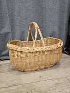 Side view of the oval handwoven willow market basket with two handles, photographed on a wooden floor against a grey curtain