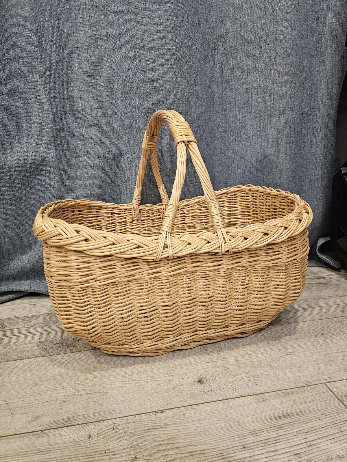 Side view of the oval handwoven willow market basket with two handles, photographed on a wooden floor against a grey curtain