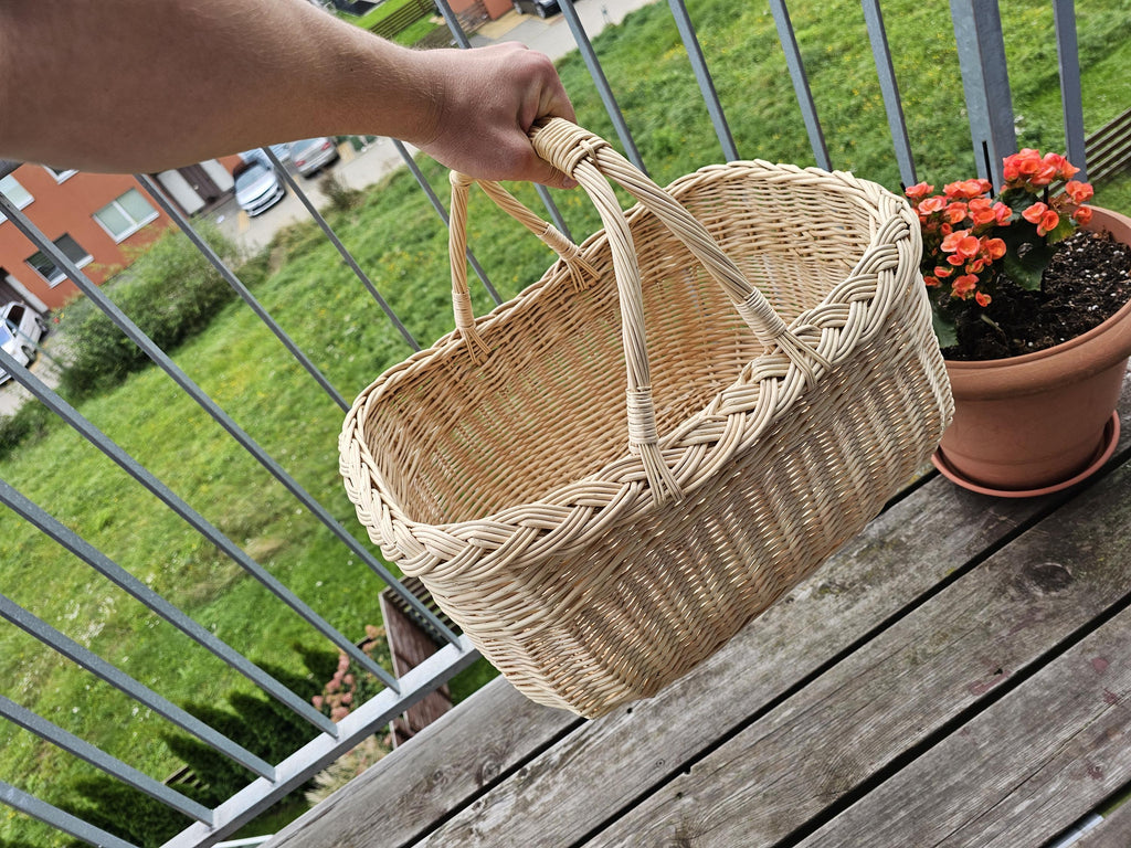 Hand carrying the large oval wicker market basket by its two joined handles on a balcony, showing its size and depth