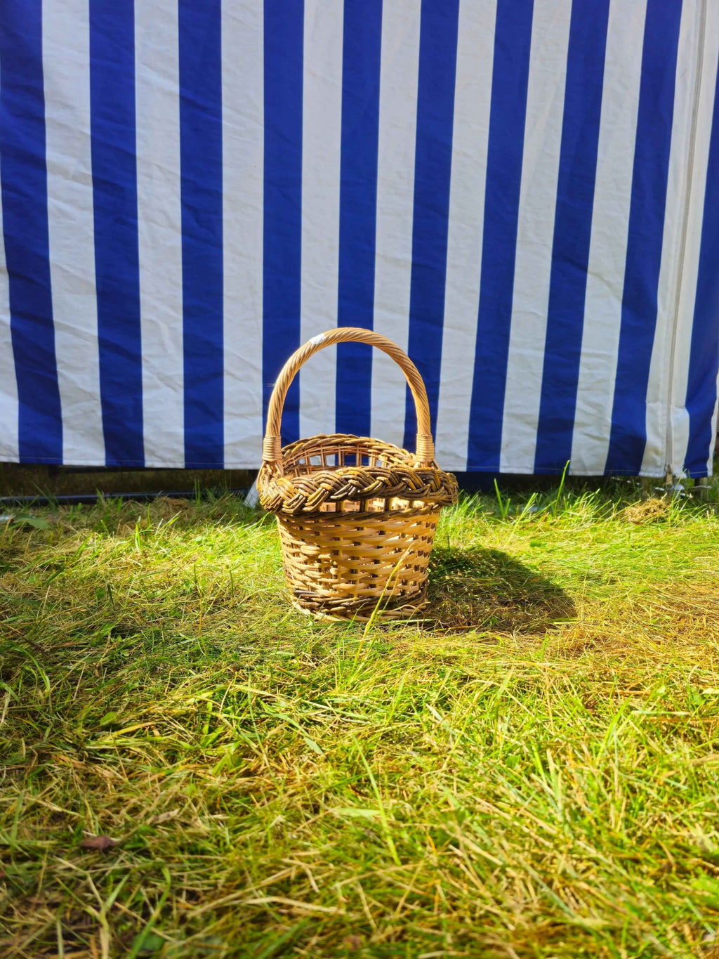 Front view of the small oval willow bread basket with handle on the grass, showing its size against a striped tent.