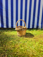 Front view of the small oval willow bread basket with handle on the grass, showing its size against a striped tent.