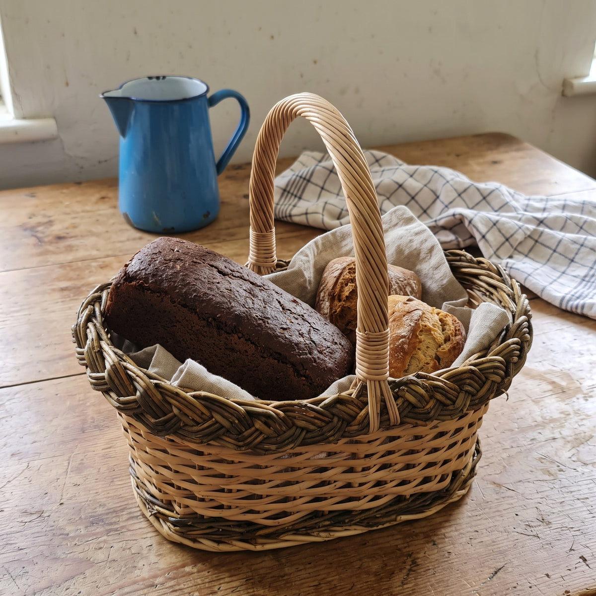 Small oval wicker bread basket with handle holding a dark rye loaf and scones on a linen cloth on a farmhouse kitchen table.