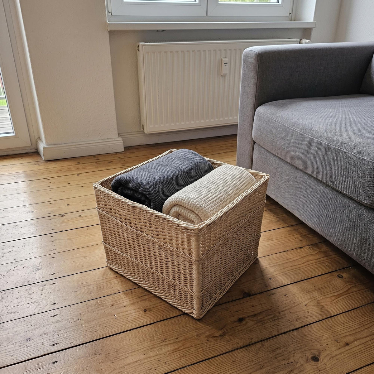 Large willow storage basket with folded blankets on a wooden floor next to a grey sofa