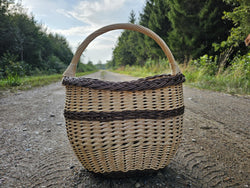 Tiefer ovaler Weidenkorb mit hohem, geschwungenem Henkel und Zopfrand auf einem Schotterweg im Wald