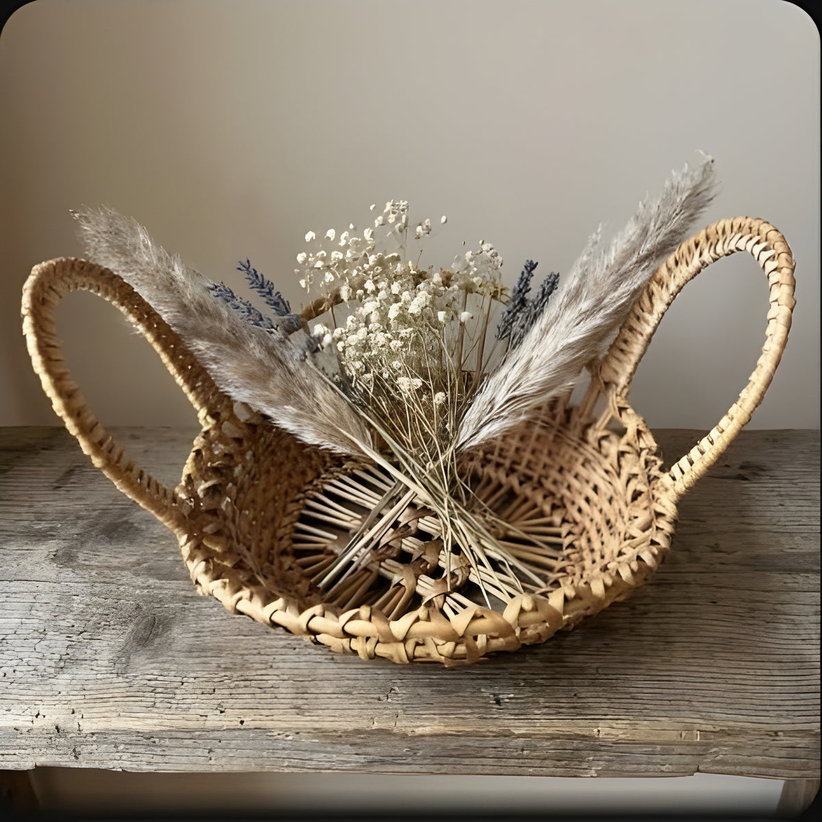 Open-weave wicker bowl with two looped handles holding dried pampas grass, gypsophila and lavender on a rustic table