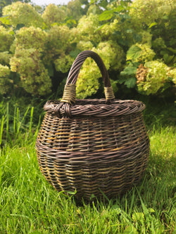 Round willow gathering basket with a thick braided rim and an arched handle bound at the base, standing on grass by hydrangea bushes