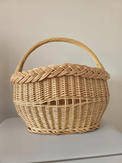 Small handwoven willow basket with a thick braided rim, an arched handle and an open-weave band, photographed against a plain wall