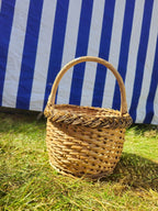Small round wicker basket with an open-weave side wall, solid circular base, arched handle and braided rim, on grass before a striped awning.
