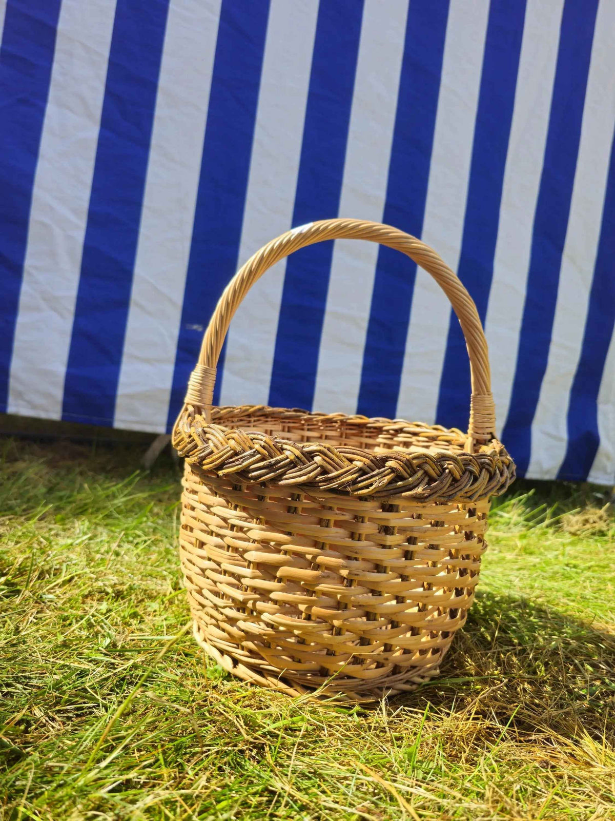 Small round wicker basket with an open-weave side wall, solid circular base, arched handle and braided rim, on grass before a striped awning.