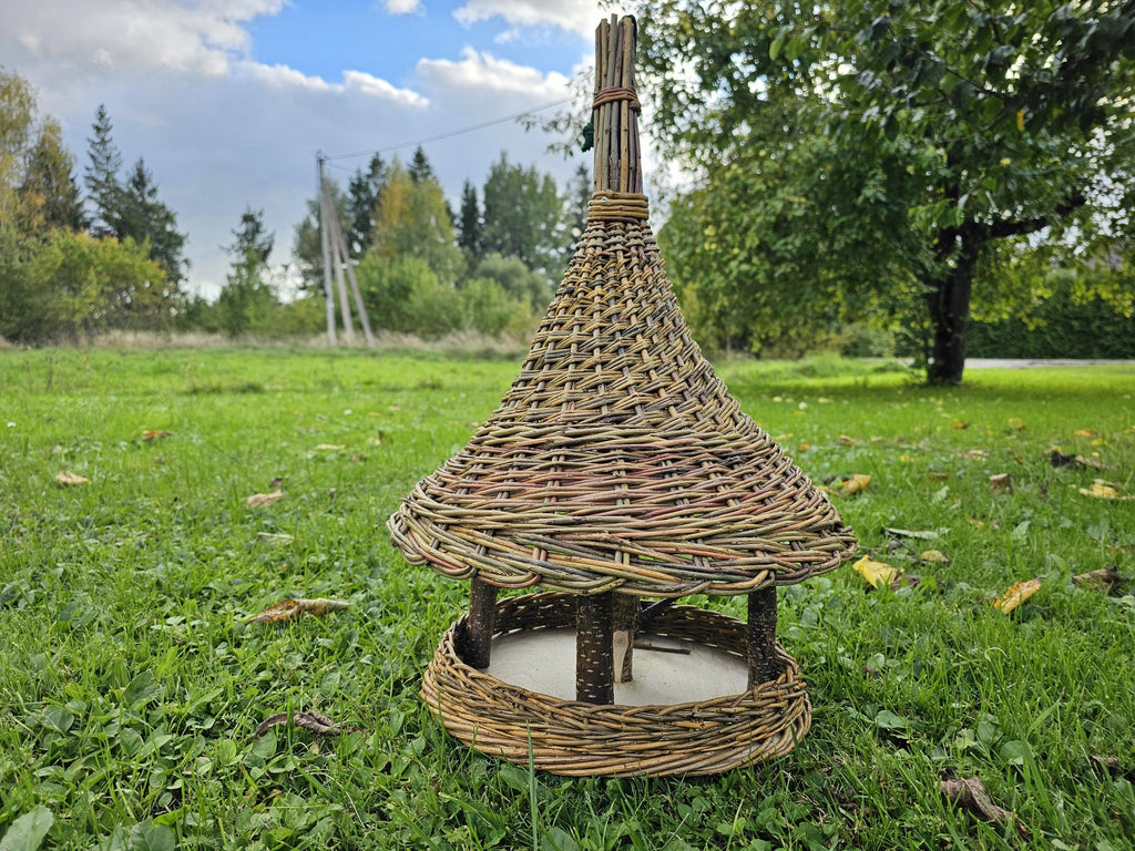 Tall willow bird feeder with bell-shaped woven roof, branch posts and round seed platform standing on a lawn