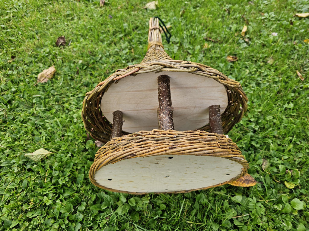 The willow bird feeder laid on its side on grass, showing the round wooden platform, branch posts and roof underside