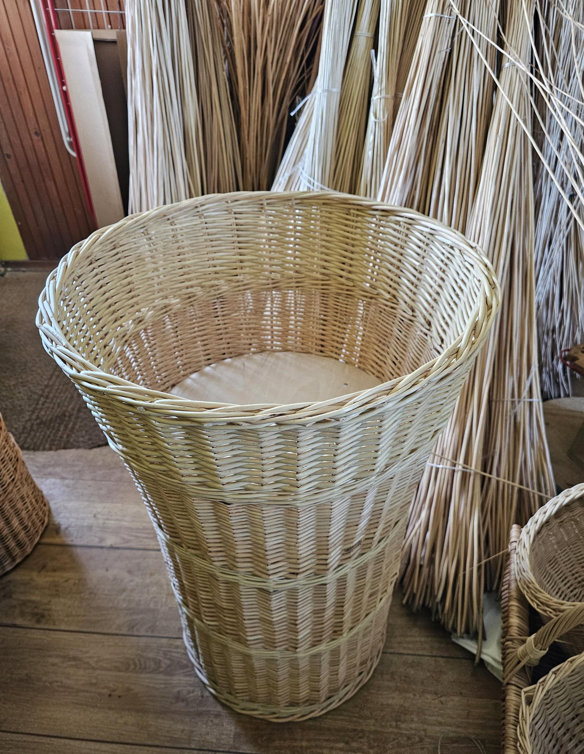 Tall conical wicker display basket in the workshop, tapering from a wide open top to a narrower base with a flat circular bottom