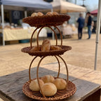 Three-tier handwoven wicker bread display stand with buns and rolls on a wooden table at an outdoor market stall