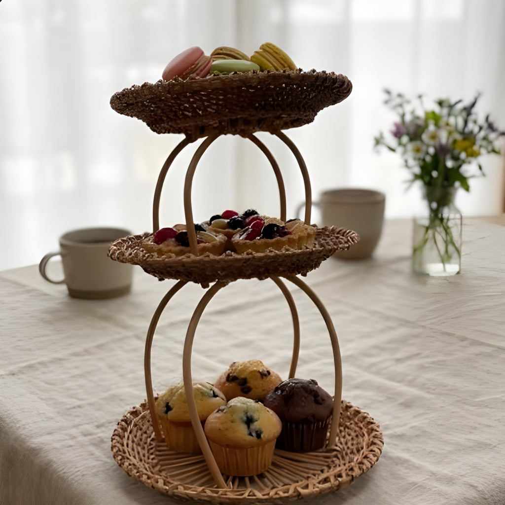 Three-tier handwoven willow display stand with macarons, berry tarts and muffins on a linen-covered table with mugs and wildflowers