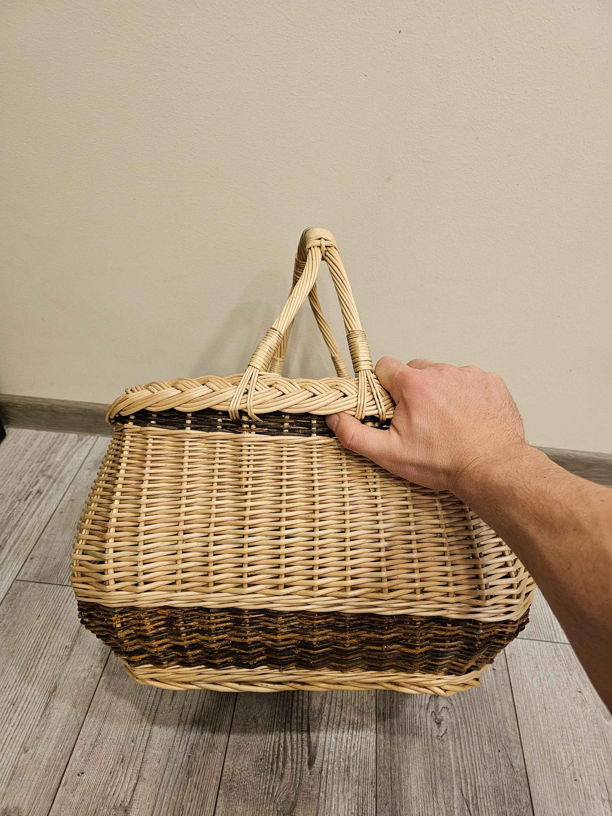 Scale shot of a handwoven willow harvest basket held by its twisted handles, a hand resting on the rim to show its generous size.