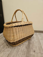 Three-quarter view of a handwoven willow harvest basket with its heavy braided edge and paired twisted handles rising above the woven body.