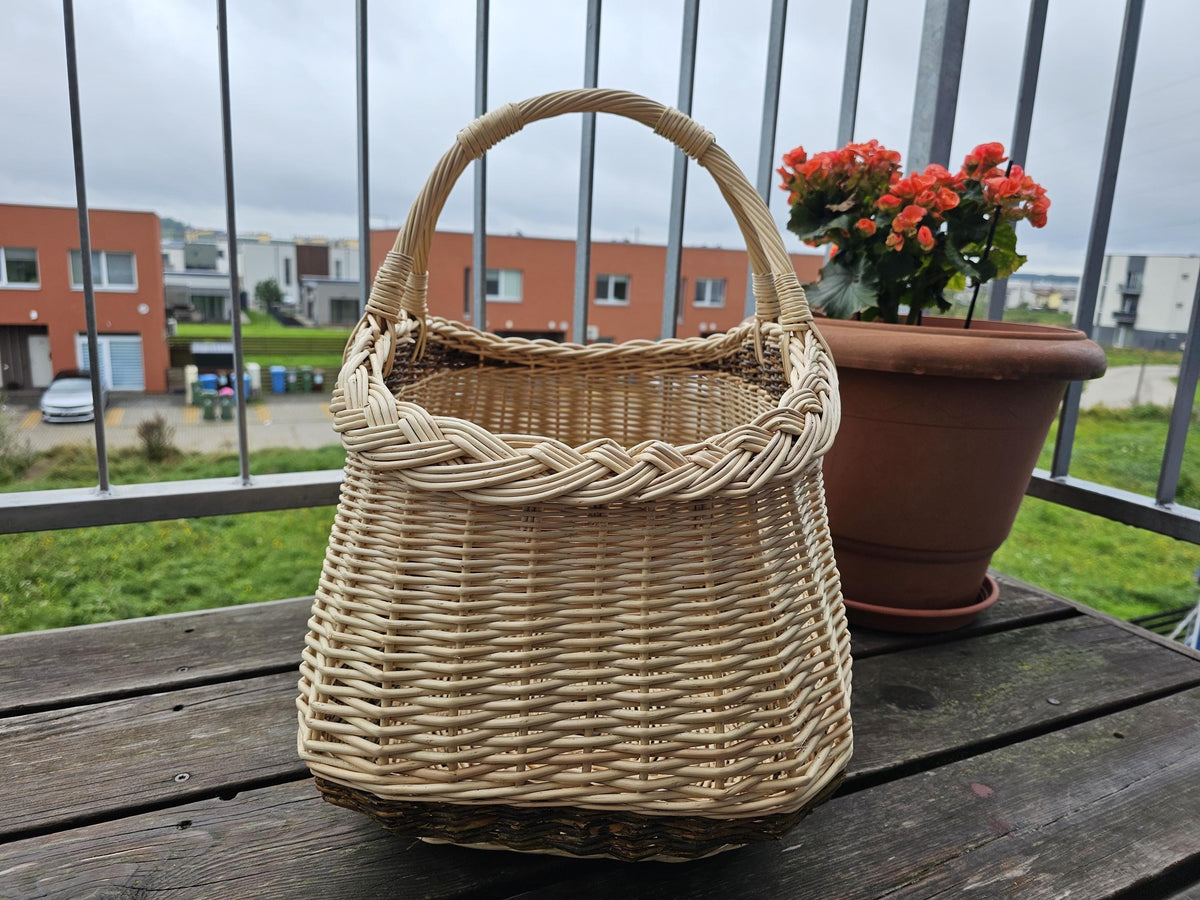 End view of the two-handled willow foraging basket showing how it widens towards the braided rim, beside a potted plant.