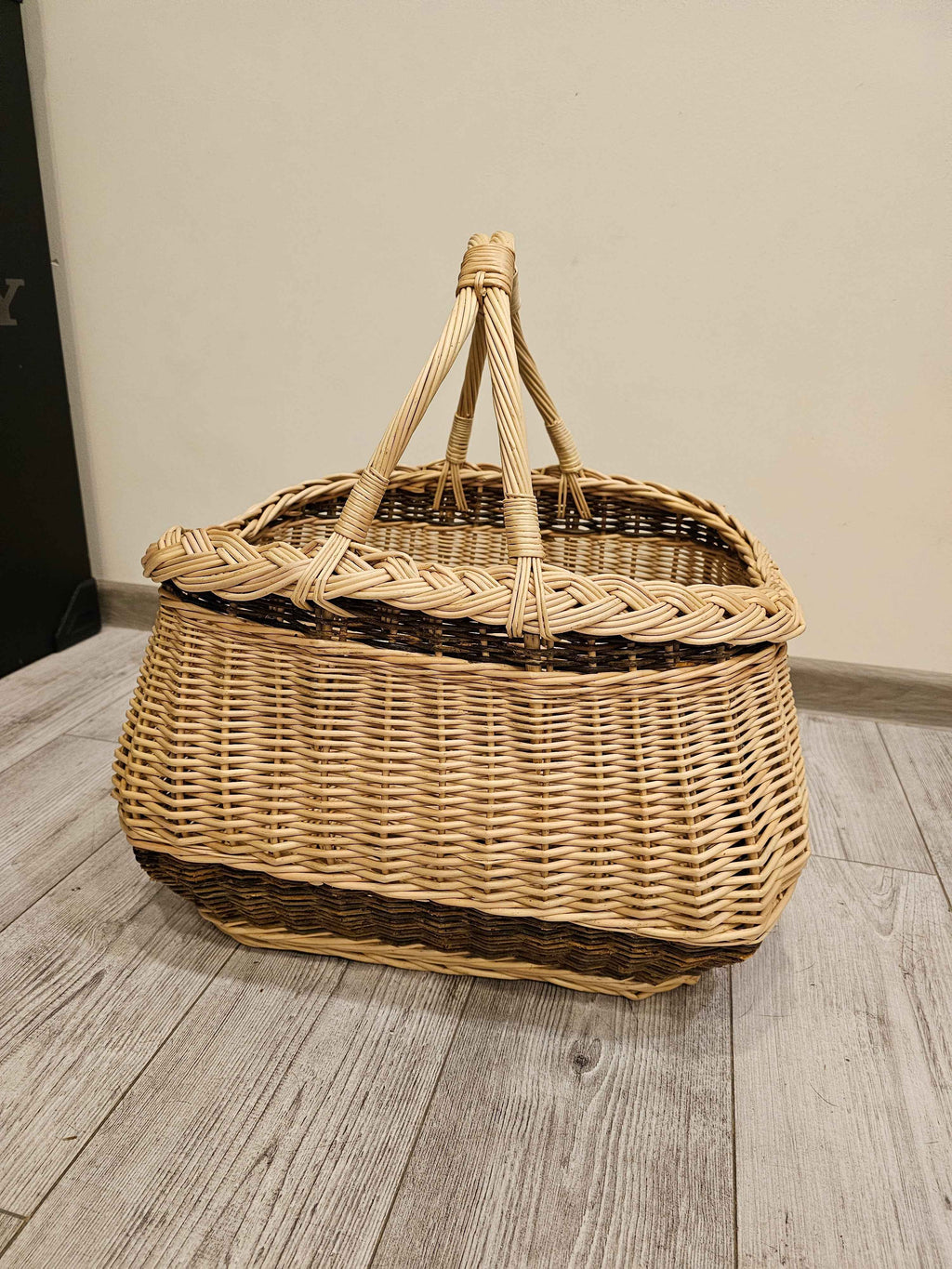 Handwoven wicker basket with a thick braided edge and rope-twisted handles bound together at the top, photographed on a grey wood floor.