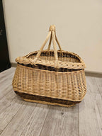 Handwoven wicker basket with a thick braided edge and rope-twisted handles bound together at the top, photographed on a grey wood floor.