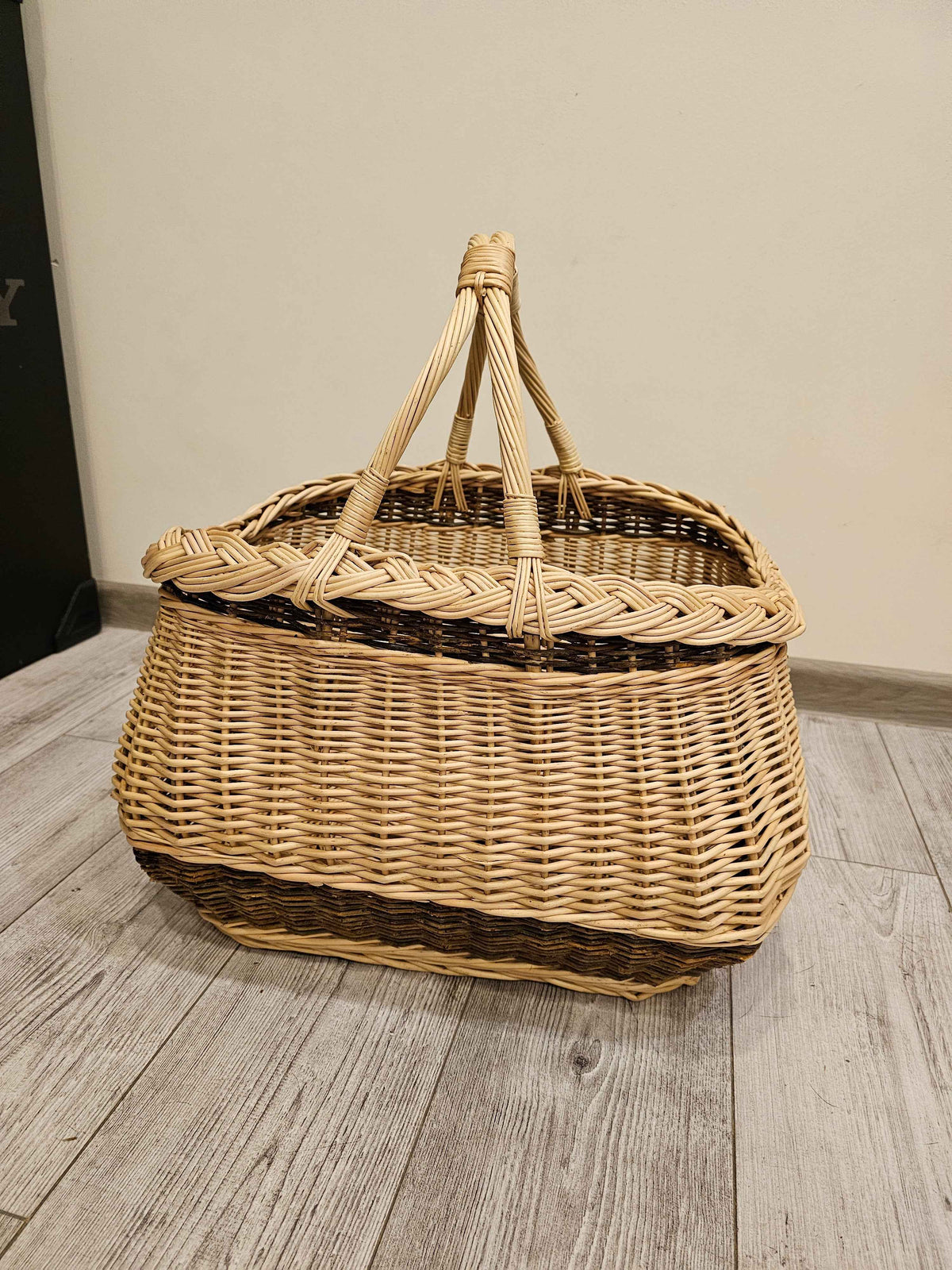 Handwoven wicker basket with a thick braided edge and rope-twisted handles bound together at the top, photographed on a grey wood floor.