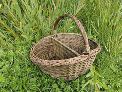 Handwoven willow basket with a sturdy arched handle and a central divider, its tight textured weave shown on long grass