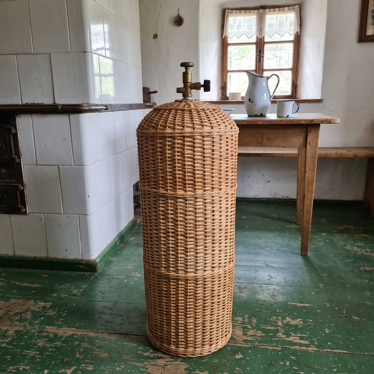 Tall handwoven willow gas cylinder cover with a domed top and valve opening, standing on a green painted floor in a country kitchen