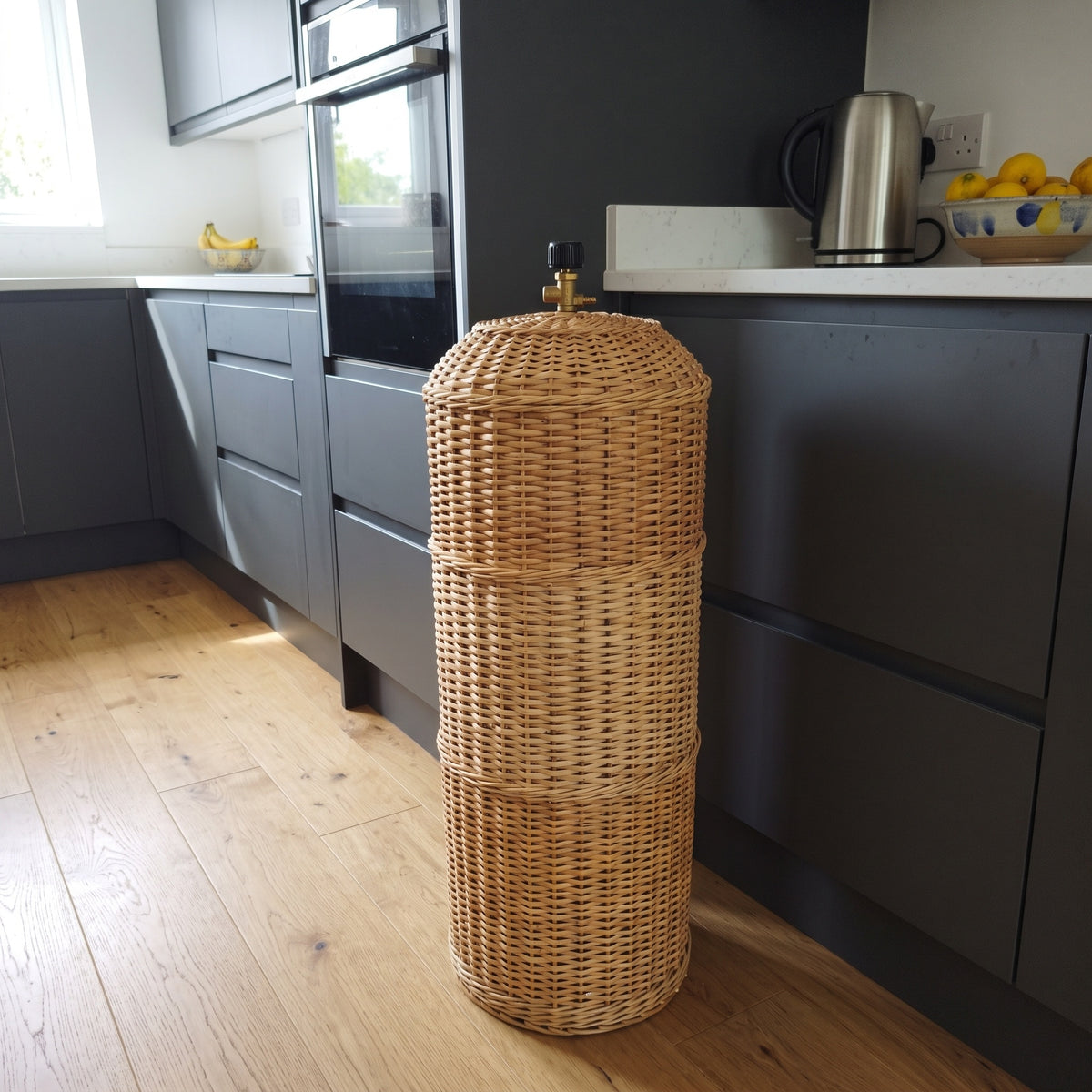Handwoven willow gas bottle cover standing beside modern dark kitchen cabinets on an oak floor, hiding a gas cylinder in the kitchen