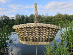 Handwoven oval willow gathering basket seen head on above a calm lake, its braided rim and tall willow handle set against a blue sky