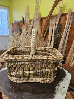 Rectangular willow tote basket with a tall twisted handle and woven bands around the body, shown against bundles of dried willow