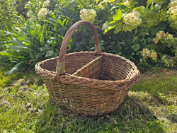 Oval wicker picnic basket with a single arched handle and centre divider on a garden lawn in front of hydrangeas.