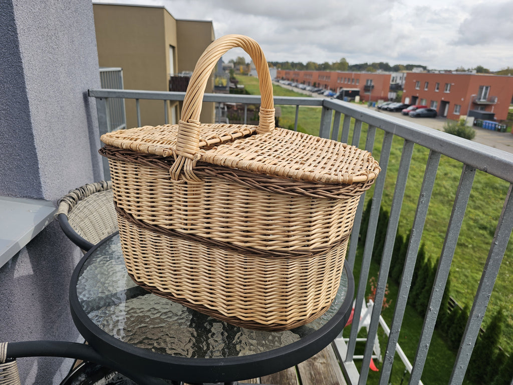 Oval wicker picnic basket with closed twin lids, a centre handle and a woven band around the body, standing on a glass table on a city balcony