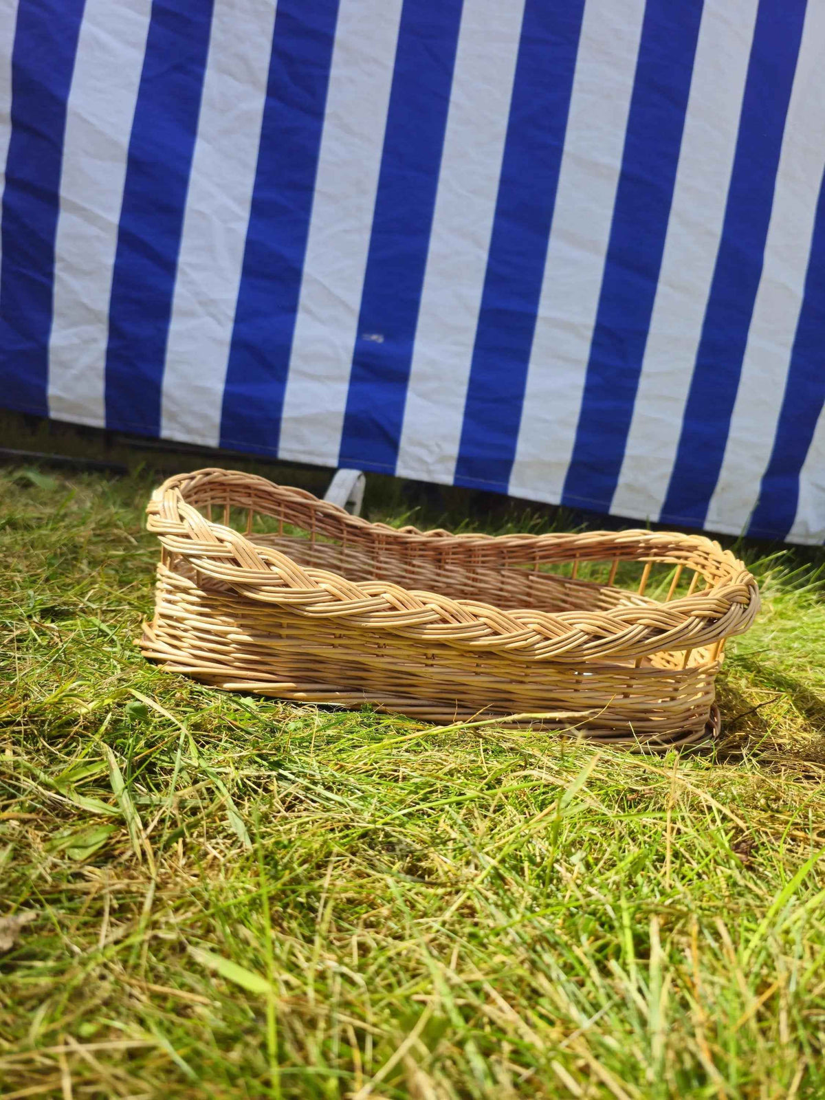 Small wicker serving tray with braided rim and raised open-weave ends resting on sunlit grass by a striped market tent