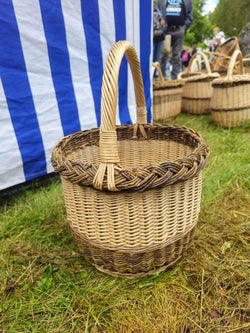 Tiefer runder Weidenkorb mit Zopfrand und Bügelhenkel im Gras auf einem Handwerkermarkt, dahinter ein gestreifter Stand und Körbe